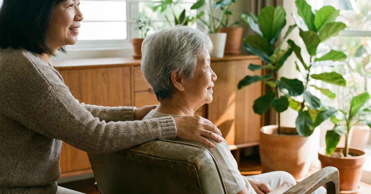 A carer rests a reassuring hand on an elderly woman's shoulder in a bright Malaysian home